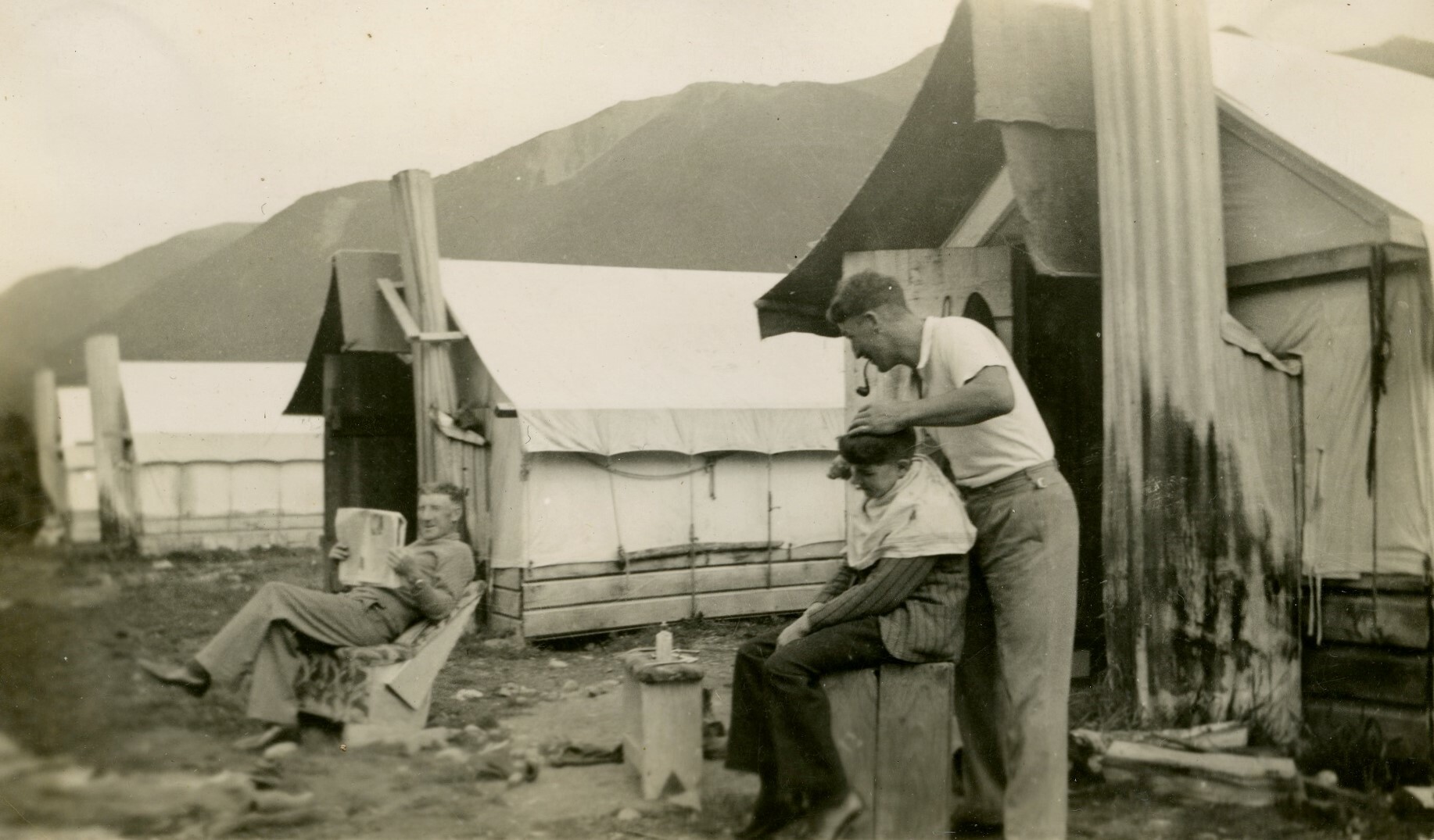 A man cuts a boys hair at the Public Works Department Camp. Roy Murphy is in the background reading a newspaper. Canterbury Museum 2019.13.28. No known copyright restrictions